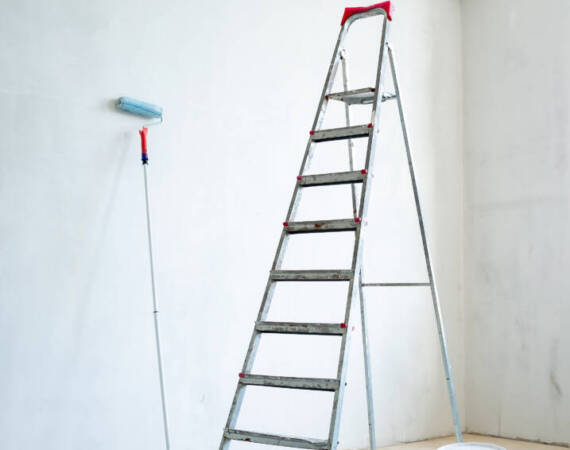 Stepladder, a closed plastic bucket of paint, and a paint roller are found in the room, in natural light from the window. Preparation for repair.